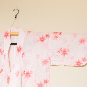 May include: A white kimono with a pink floral pattern, hanging from a wooden hanger. The kimono features a repeating design of pink cherry blossoms. The garment is displayed against a neutral background.