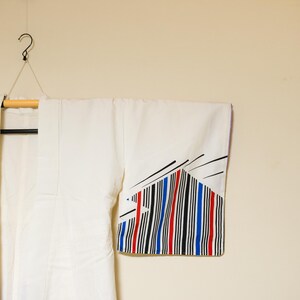 May include: White kimono hanging on a wooden hanger. The sleeve features a geometric design with black, red, and blue vertical stripes. The garment is suspended against a neutral-toned wall.