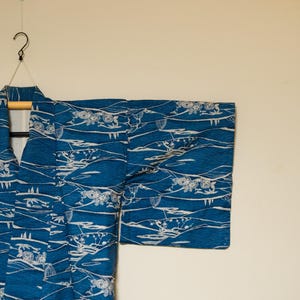 Op de afbeelding: Een blauwe kimono met een wit patroon van gestileerde golven en bloemen. De kimono hangt aan een hanger.