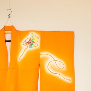 Op de afbeelding: Fel oranje kimono met een decoratief ontwerp aan de rechterkant. Het ontwerp heeft een gele vliegerachtige vorm met kleurrijke bloemaccenten en een vloeiende witte lijn. De kimono hangt aan een zwarte hanger.