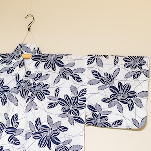 May include: White kimono with a navy blue floral pattern hanging from a wooden hanger. The floral design features stylized flowers and leaves. The garment is made of a lightweight fabric, suitable for warm weather. The kimono is displayed against a neutral background.