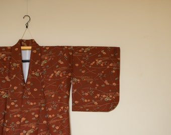May include: A rust-colored kimono with a floral pattern, hanging from a black hook. The kimono features a repeating design of flowers and flowing lines in shades of orange, green, and cream. The garment is made of a soft fabric.