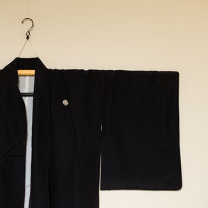 May include: A black kimono hanging on a wooden hanger. The kimono has a simple design and is made of a smooth fabric.