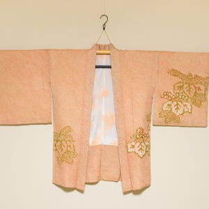 May include: A pale pink Japanese kimono with gold embroidered floral designs. The kimono is hanging on a wooden hanger.