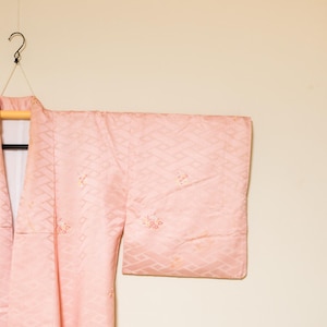 Puede incluir: Un kimono rosa claro con un patrón geométrico, colgado de una percha negra. El kimono está hecho de una tela suave y fluida y tiene mangas anchas. El fondo es de color beige claro.