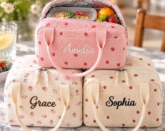 Peut inclure: Trois sacs à déjeuner rectangulaires en velours côtelé avec des motifs de fraises. Le sac du haut est rose avec le prénom Amelia brodé en blanc. Les deux sacs du bas sont crème avec les prénoms Grace et Sophia brodés en marron.
