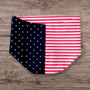 Può includere: Un bandana patriottico per animali domestici con una sezione blu navy con croci bianche e una sezione a righe rosse e bianche. Il bandana è piegato e poggia su una superficie di legno.