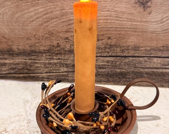 Puede incluir: Una vela cónica naranja que funciona con pilas y tiene una llama LED brillante se encuentra en un portavelas de metal marrón. La base de la vela está rodeada por un anillo decorativo de ramas marrones con pequeñas bayas negras y naranjas.