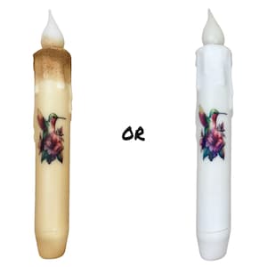 May include: Two LED flameless candles with a hummingbird and flower design. The candle on the left is a cream color with a brown drip effect. The candle on the right is white.