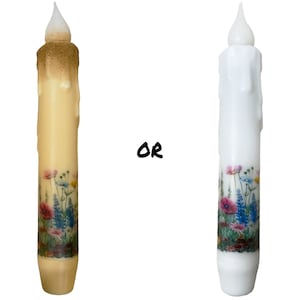 May include: Two decorative candles with a floral design. The candle on the left is a light yellow color and the candle on the right is white. Both candles have a white wax drip effect and a white wick.