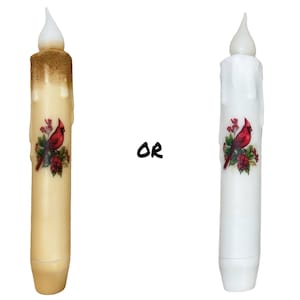 May include: Two battery-operated LED candles with a cardinal bird and holly design. The candle on the left is cream colored and the candle on the right is white.