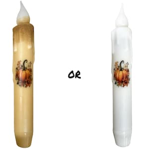 以下が含まれることがあります： かぼちゃと秋の葉っぱのデザインが施された、装飾的な円柱型のろうそくが2本。左側のろうそくはクリーム色、右側のろうそくは白です。