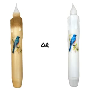 May include: Two LED flameless candles with a bluebird design. The candle on the left is beige and the candle on the right is white.