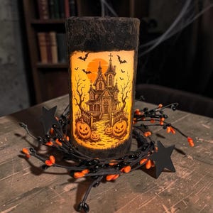 May include: A cylindrical pillar candle with a dark, textured exterior featuring a glowing orange insert. The insert displays a black illustration of a haunted house, bare trees, flying bats, and jack-o'-lanterns. A black star-accented garland surrounds the base.