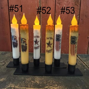 Op de afbeelding: Zes LED-kaarsen met patriottische ontwerpen. De kaarsen hebben een flikkerend vlameffect en zijn versierd met Amerikaanse vlaggen, adelaars en de tekst "God Bless America" en "We the People". Ze staan op een zwarte metalen basis.