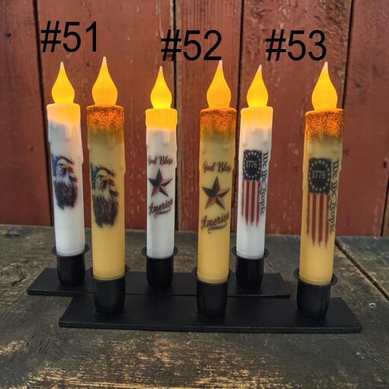 Op de afbeelding: Zes LED-kaarsen met patriottische ontwerpen. De kaarsen hebben een flikkerend vlameffect en zijn versierd met Amerikaanse vlaggen, adelaars en de tekst "God Bless America" en "We the People". Ze staan op een zwarte metalen basis.