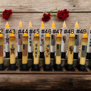 May include: A collection of battery-operated LED candles with patriotic designs. Each candle has a flickering yellow light and is decorated with American flags, stars, and the word "HOME". The candles are arranged on a wooden surface.