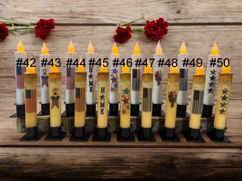 Op de afbeelding: Een verzameling op batterijen werkende LED-kaarsen met patriottische ontwerpen. Elke kaars heeft een flikkerend geel licht en is versierd met Amerikaanse vlaggen, sterren en het woord "HOME". De kaarsen zijn gerangschikt op een houten oppervlak.