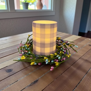May include: A glowing, cylindrical candle with a gray and white checkered pattern. The candle is surrounded by a decorative wreath of green stems and colorful, small, round berries. The candle and wreath sit on a wooden table.