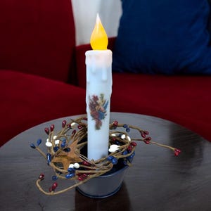 Op de afbeelding: Een batterijkaars met een flikkerende gele vlam staat in een kleine, donkerblauwe metalen emmer. De kaars is versierd met een bloemenontwerp en omgeven door een slinger van rode, witte en blauwe bessen.