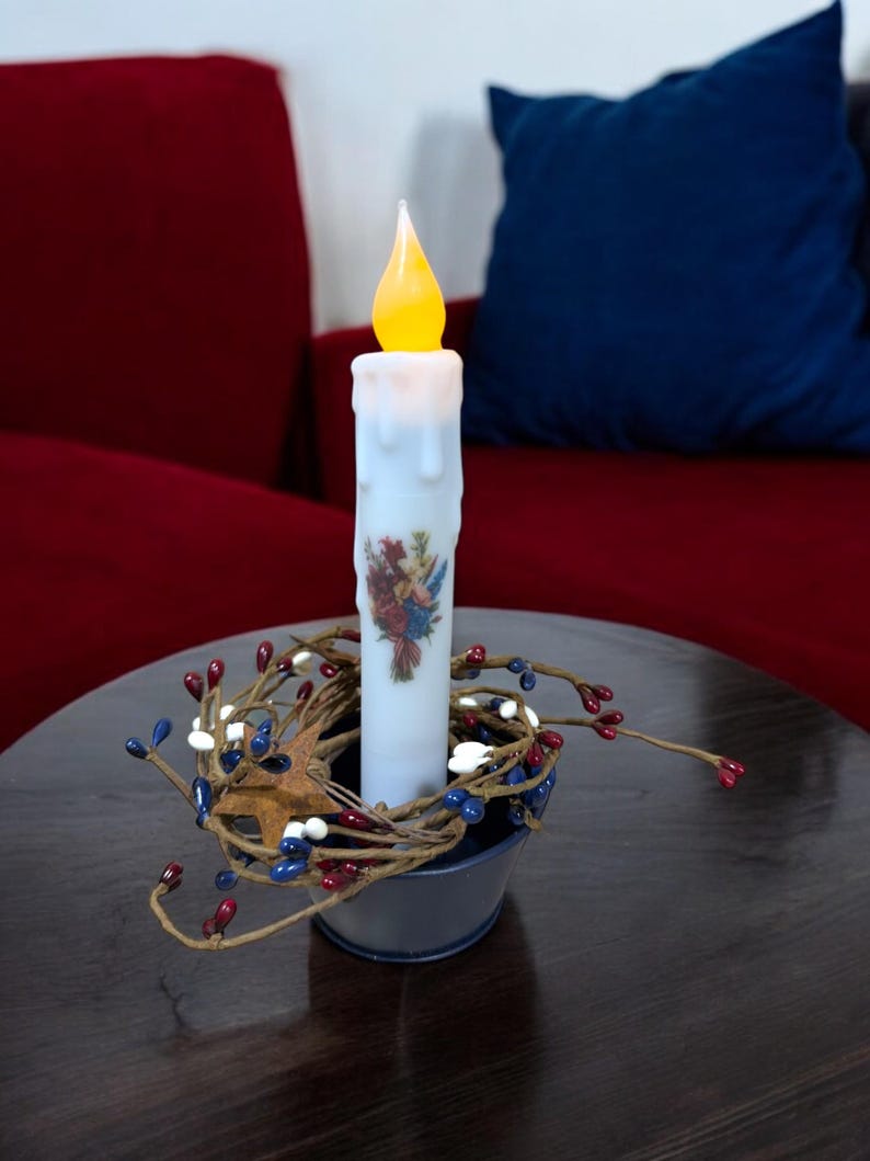 Op de afbeelding: Een batterijkaars met een flikkerende gele vlam staat in een kleine, donkerblauwe metalen emmer. De kaars is versierd met een bloemenontwerp en omgeven door een slinger van rode, witte en blauwe bessen.