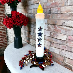 Op de afbeelding: Een witte LED-kaars met een druipend wax-effect en ster-decals, geplaatst op een zwarte basis met een rode, witte en blauwe bessenkrans. Rode bloemen in zwarte vazen staan op de achtergrond. De kaars is ongeveer 25 cm hoog.