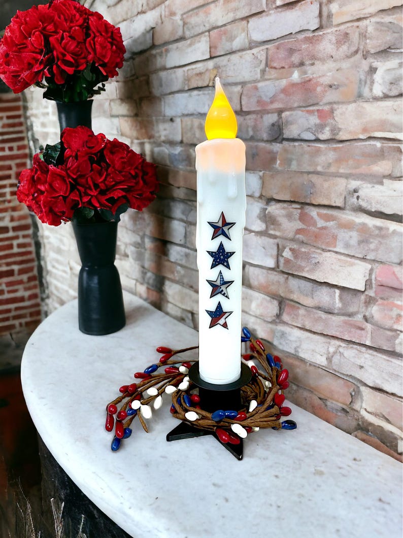 Op de afbeelding: Een witte LED-kaars met een druipend wax-effect en ster-decals, geplaatst op een zwarte basis met een rode, witte en blauwe bessenkrans. Rode bloemen in zwarte vazen staan op de achtergrond. De kaars is ongeveer 25 cm hoog.