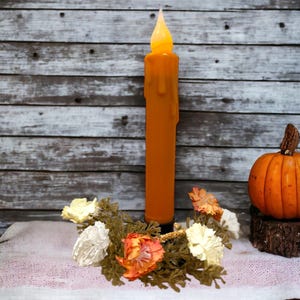 May include: A tall, orange, drippy candle with a flickering flame sits on a wooden surface with a white lace runner.  The candle is surrounded by a wreath of white and orange flowers.