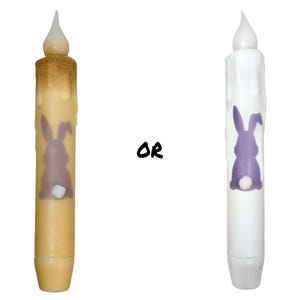 May include: Two battery-operated Easter candles. One is beige with a brown top and a purple bunny design. The other is white with a purple bunny design. The word "OR" is between the candles.