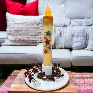 Op de afbeelding: Een batterijkaars met een flikkerende vlam, versierd met rode, witte en blauwe sterren. De kaars staat op een wit bord met een bessenkrans, geplaatst op een houten plank. De kaars is ongeveer 20 cm hoog.