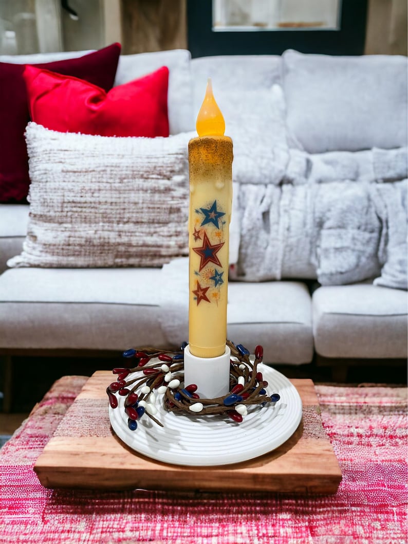 Op de afbeelding: Een batterijkaars met een flikkerende vlam, versierd met rode, witte en blauwe sterren. De kaars staat op een wit bord met een bessenkrans, geplaatst op een houten plank. De kaars is ongeveer 20 cm hoog.