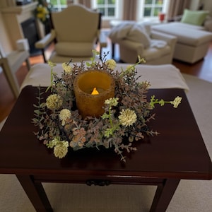 May include: A decorative candle holder with a faux flame, surrounded by a wreath of greenery and small white flowers. The candle holder is on a dark brown wooden table, creating a warm and inviting ambiance.