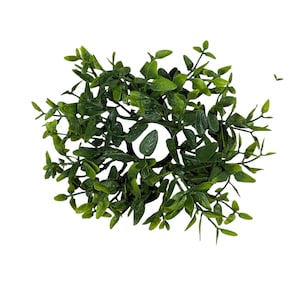 May include: A round, artificial greenery candle ring. The ring is composed of dark green leaves and small branches. The leaves are a mix of shapes and sizes, creating a natural, textured appearance. The candle ring is designed to hold a candle.