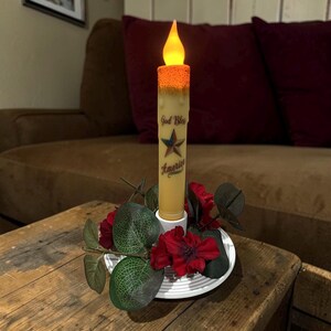 Op de afbeelding: Een batterijkaars met een flikkerende amberkleurige vlam. De kaars is versierd met de tekst "God Bless America" en een ster. Hij staat op een wit bord omringd door rode bloemen en groene bladeren, geplaatst op een houten tafel.