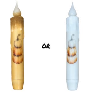 May include: Two white pillar candles with a stack of three pumpkins on each. The pumpkins are painted in shades of orange and brown. The candles are decorated with a white wax drip effect.  The text "OR" is written in black between the candles.