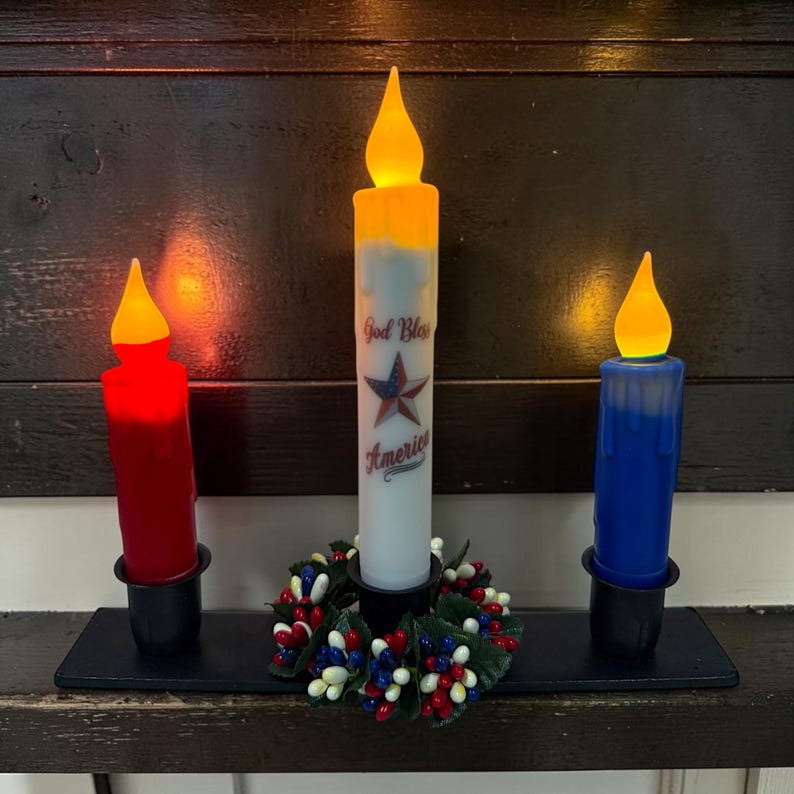 Op de afbeelding: Drie LED-kaarsen in rood, wit en blauw, elk in een zwarte houder, met een patriottisch thema. De witte kaars heeft de tekst "God Bless America" en een sterrenafbeelding. Een kleine krans met rode, witte en blauwe bessen staat aan de voet van de middelste kaars.