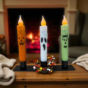 May include: Three Halloween-themed LED candles in orange, white, and green, each with a different face design. The candles sit in black holders on a black base, with a small wreath of candy corn and berries. The flames are a warm yellow.