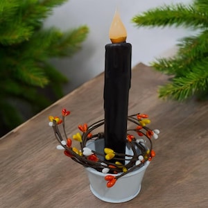 May include: A black pillar candle with a flickering LED flame sits in a white metal bucket. The bucket is decorated with a wreath of orange, yellow, and white berries.