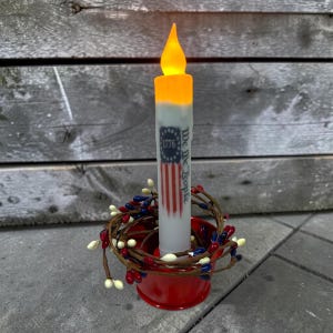 Op de afbeelding: Een batterijgevoede kaars met een patriottisch ontwerp. De kaars heeft een gedrukte Amerikaanse vlag en de tekst "We the People 1776". Hij staat in een rode metalen houder en is omgeven door een bessenkrans.