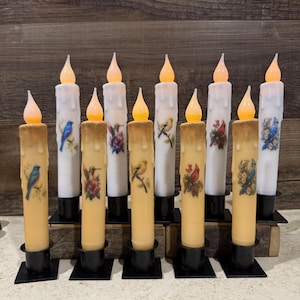 May include: Ten ivory-colored pillar candles with a variety of bird designs. The candles are lit and have a warm glow. The candles are on a wooden surface.