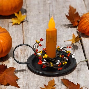 May include: An orange candle sits in a black metal candle holder, surrounded by a berry garland. The candle is on a wooden surface with pumpkins and fall leaves, creating a seasonal autumn theme.