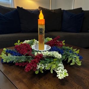Op de afbeelding: Een patriottische kaars met een flikkerende LED-vlam, met een ontwerp van de Amerikaanse vlag en de tekst "We the People". De kaars staat in een wit schaaltje, omringd door een krans van rode, witte en blauwe bloemaccenten.