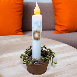 Może przedstawiać: Biała świeca w kształcie kolumny z wzorem wieńca słonecznikowego, zapalona migoczącym płomieniem, stoi w brązowym metalowym pojemniku otoczonym zielonym wieńcem.