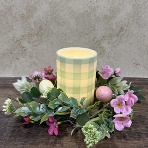May include: A cylindrical candle with a green and yellow checkered pattern, surrounded by a wreath of pink flowers, green leaves, and decorative Easter eggs. The candle is lit, creating a warm glow, and sits on a dark wooden surface.