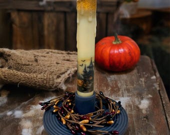 May include: A battery-operated candle with a flickering flame and a spooky house design. The candle is placed in a blue ceramic candle holder with a decorative berry garland.