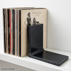 May include: A black metal L-shaped bookend next to a stack of vinyl records. The album cover visible features the Fleetwood Mac Rumours album. The bookend is on a white shelf.