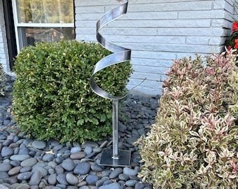 May include: A modern metal sculpture in the shape of a spiral, standing on a square base. The sculpture is made of silver metal and is placed in a garden setting with gray stones and green bushes.