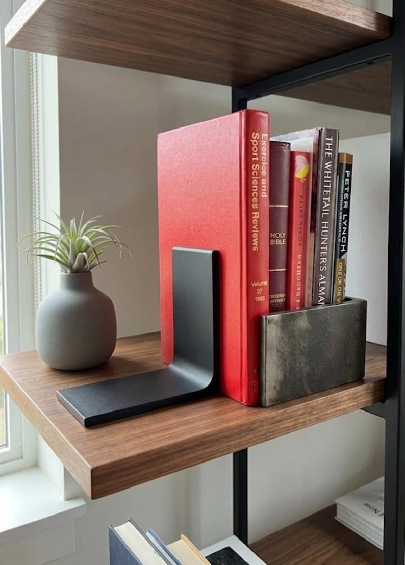 May include: A modern bookshelf with a wooden shelf holding books and decorative items. A red book is held in place by a black metal bookend. A small gray vase with a plant sits to the left of the books. The shelf is part of a black metal frame.