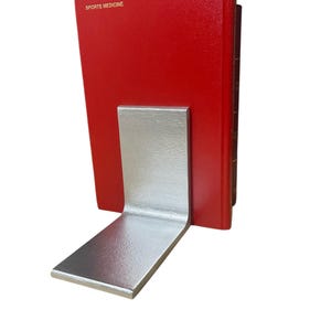 May include: A silver metal bookend supporting a red book with the words "SPORTS MEDICINE" printed on the cover. The bookend has a simple, L-shaped design, with a flat base and a vertical support.