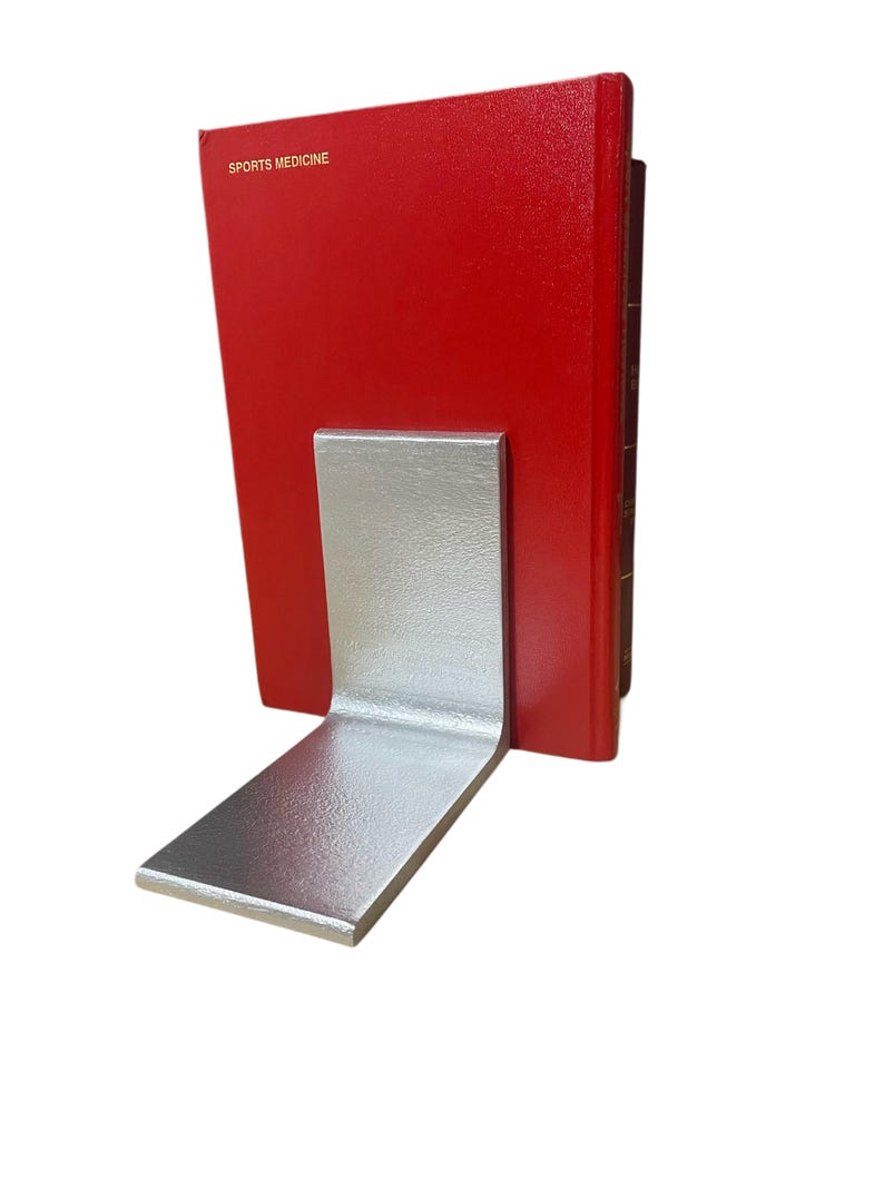 May include: A silver metal bookend supporting a red book with the words "SPORTS MEDICINE" printed on the cover. The bookend has a simple, L-shaped design, with a flat base and a vertical support.